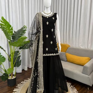 May include: A black sleeveless embroidered outfit with a matching skirt and a sheer, embellished shawl. The outfit is displayed on a mannequin, showcasing the intricate silver embroidery and trim. The ensemble is set against a neutral backdrop.