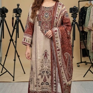 Puede incluir: Conjunto salwar kameez estampado en beige y granate. El atuendo incluye una túnica larga, pantalones a juego y un chal. La túnica y el chal presentan intrincados estampados florales y de cachemira. La modelo lleva tacones negros.