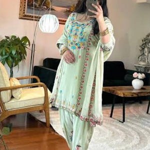 Puede incluir: Conjunto salwar kameez bordado en verde claro. El atuendo presenta intrincados bordados florales en tonos azules, rosas y amarillos. El conjunto incluye una túnica, pantalones a juego y una bufanda.