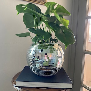 May include: A large green monstera plant in a silver disco ball planter. The planter is sitting on top of a black book on a wooden table.