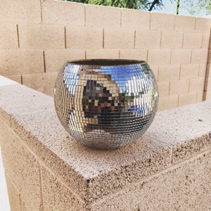 May include: A silver disco ball planter with a mirrored surface. The planter is sitting on a light brown textured wall.