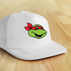 May include: White trucker hat featuring a green cartoon turtle face with a red headband. The turtle has a smiling expression. The hat has a white mesh back and a curved brim.