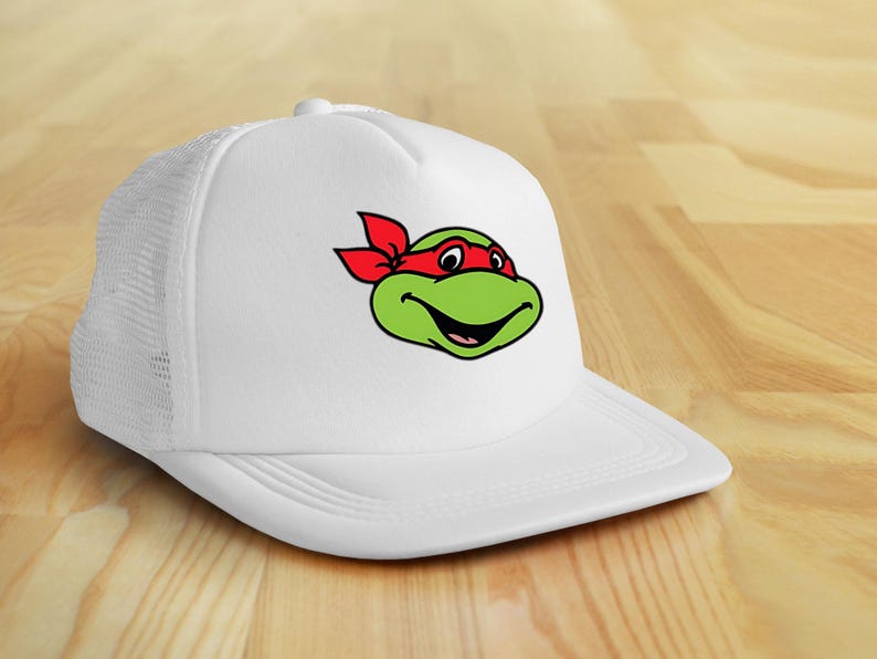 May include: White trucker hat featuring a green cartoon turtle face with a red headband. The turtle has a smiling expression. The hat has a white mesh back and a curved brim.
