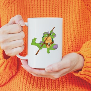 May include: White ceramic mug featuring a cartoon image of Donatello from Teenage Mutant Ninja Turtles. Donatello is depicted in green, purple, and brown, holding his bo staff. The mug has a standard handle and a glossy finish.