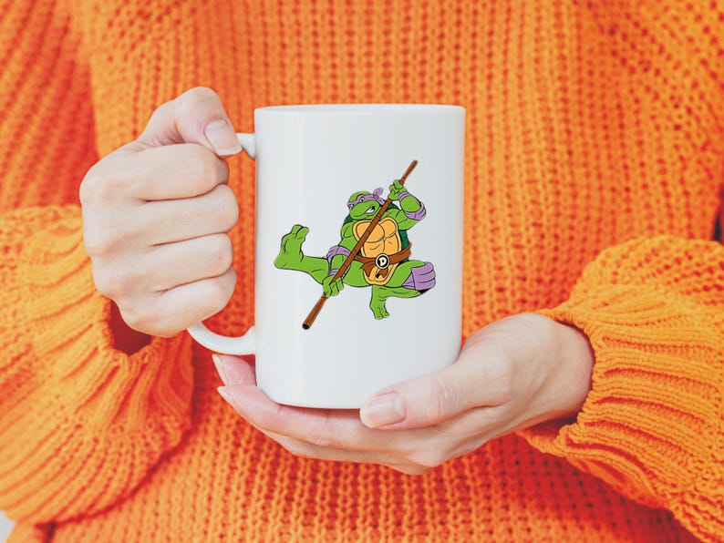 May include: White ceramic mug featuring a cartoon image of Donatello from Teenage Mutant Ninja Turtles. Donatello is depicted in green, purple, and brown, holding his bo staff. The mug has a standard handle and a glossy finish.