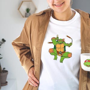 May include: White t-shirt featuring a green cartoon ninja turtle character with orange accents, holding weapons. A matching mug with a turtle face is also visible. The shirt and mug are part of a themed collection.