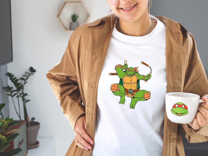 May include: White t-shirt featuring a green cartoon ninja turtle character with orange accents, holding weapons. A matching mug with a turtle face is also visible. The shirt and mug are part of a themed collection.
