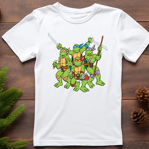 May include: White t-shirt featuring a graphic of the Teenage Mutant Ninja Turtles. The turtles are green with colored masks and holding weapons. The design is centered on the shirt.