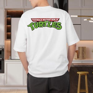 May include: White t-shirt featuring the text "TEENAGE MUTANT NINJA TURTLES" in green and red. The shirt has short sleeves and a relaxed fit. The design is on the back of the shirt.