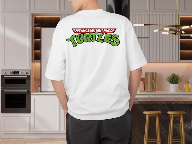 May include: White t-shirt featuring the text "TEENAGE MUTANT NINJA TURTLES" in green and red. The shirt has short sleeves and a relaxed fit. The design is on the back of the shirt.