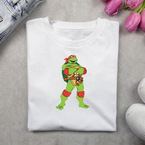 May include: White t-shirt featuring a cartoon image of Raphael from Teenage Mutant Ninja Turtles. The green turtle character is wearing a red mask, arm and knee bands, and a brown belt. The shirt is folded on a gray surface.