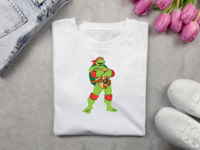 May include: White t-shirt featuring a cartoon image of Raphael from Teenage Mutant Ninja Turtles. The green turtle character is wearing a red mask, arm and knee bands, and a brown belt. The shirt is folded on a gray surface.