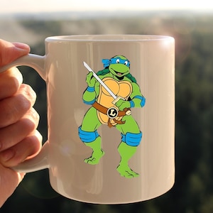 May include: White ceramic mug featuring a cartoon image of Leonardo, a Teenage Mutant Ninja Turtle, holding a sword. The character is green, with blue accents and a brown belt. The mug has a white handle.