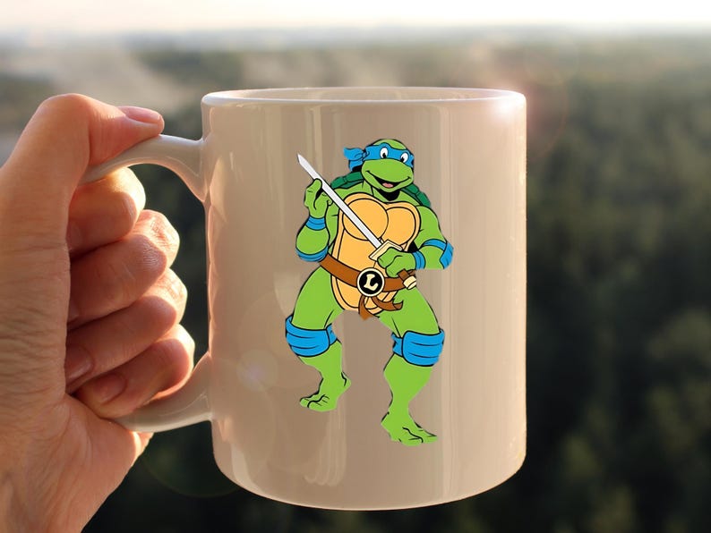 May include: White ceramic mug featuring a cartoon image of Leonardo, a Teenage Mutant Ninja Turtle, holding a sword. The character is green, with blue accents and a brown belt. The mug has a white handle.