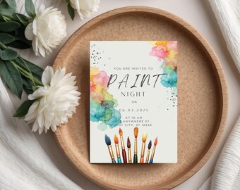 May include: A rectangular invitation card with the text "PAINT NIGHT" and watercolor splashes of pink, yellow, and blue. The card includes the date, time, and address. Paintbrushes are at the bottom. The card is on a wooden tray with white flowers.