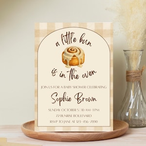 May include: Baby shower invitation featuring a cinnamon roll illustration and the phrase "a little bun is in the oven." The invitation is for Sophie Brown, including the date, time, and address. The background has a beige gingham pattern.