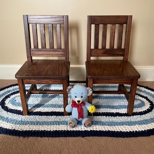 May include: Two small, wooden chairs with a dark finish stand on a blue and white oval rug. A blue crocheted teddy bear with a red bow and yellow flower sits on the rug between the chairs. The chairs have a slatted back.