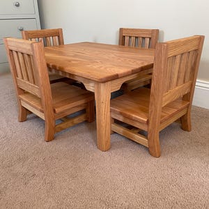 May include: A small wooden table and four matching chairs crafted from light brown wood with a visible grain. The table has a rectangular top with rounded corners and four sturdy legs. Each chair features a slatted backrest and a flat seat.