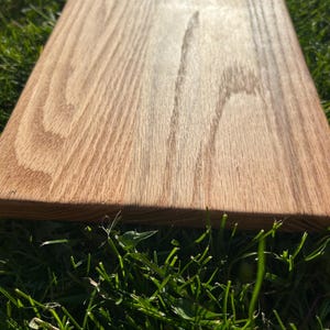May include: A rectangular wooden cutting board with a natural wood grain pattern. The board is a light brown color and appears to be made of hardwood. The board is sitting on a bed of green grass.