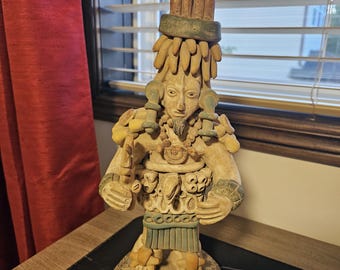 May include: A detailed, beige-colored statue of a figure with a tall headdress and intricate carvings. The statue has a beard and is adorned with various decorative elements, including a skirt and jewelry. The figure stands on a round base.