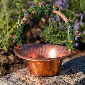 May include: A hammered copper basket with a decorative handle adorned with green moss, lavender, and small purple flowers. The basket is sitting on a rock, with a blurred background of purple flowers.