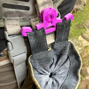 Może przedstawiać: Zbliżenie fioletowego plastikowego klipsa mocującego szarą sakiewkę do plecaka. Klips jest przymocowany do pasków plecaka i utrzymuje sakiewkę na miejscu. Sakiewka jest szara i ma zamknięcie ze sznurkiem.