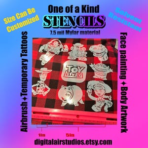 May include: A set of white Mylar stencils featuring Toy Story characters including Buzz Lightyear, Woody, Jessie, Mr. Potato Head, Rex, Hamm, and an Alien, with a Toy Story logo stencil, placed above a pink ruler showing a 12.7 cm measurement.