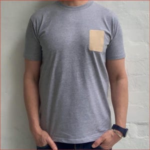 May include: Grey short-sleeved t-shirt with a square, tan pocket on the chest. The shirt has a crew neck and is made of a soft material. The person is wearing a blue watch with a silver strap.