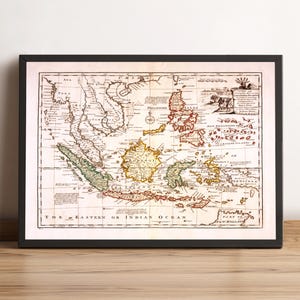 May include: Framed vintage map of Southeast Asia and surrounding islands, including the Philippines and New Guinea. The map features detailed illustrations and text, with colors highlighting different regions. The frame is black, and the map is displayed on a wooden surface.