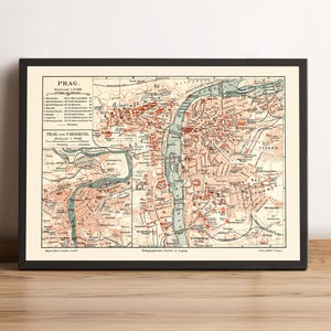 May include: Framed vintage map of Prague, featuring detailed city streets, landmarks, and the Vltava River. The map is in shades of beige, red, and blue, with text in German. The map is in a black frame and is displayed on a wooden surface.