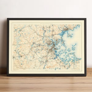 May include: Framed vintage map of the Greater Boston area, featuring detailed cartography in shades of beige, brown, and blue. The map is encased in a black frame, showcasing the intricate network of roads, rivers, and coastal features.