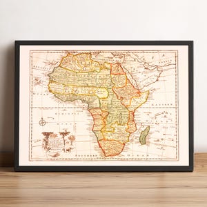 May include: A framed vintage map of Africa, featuring detailed cartography with various regions labeled. The map is in shades of beige, yellow, and red, with a black frame. The map is displayed on a wooden surface.