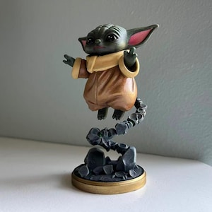 Op de afbeelding: Een beeldje van een personage uit een populaire sciencefiction-serie. De figuur is afgebeeld in een bruine outfit met een beige kraag en zweeft boven een rotsachtige basis. De figuur heeft grote oren en gebaart met zijn handen.