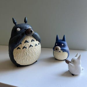 以下が含まれることがあります： アニメキャラクター、トトロの3体のフィギュア。一番大きいのは濃い青色で、クリーム色の腹を持ち、茶色の物体を持っています。中くらいのトトロは青色で、一番小さいのは白色で、どちらも似たような物体を持っています。すべて白い表面にあります。