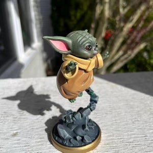 Op de afbeelding: Een beeldje van een groen alien-personage met grote oren en grote ogen, gekleed in een bruine jurk en een beige sjaal. Het beeldje zweeft in de lucht, ondersteund door een rotsachtige basis met een gouden rand. Een verzamelobject.