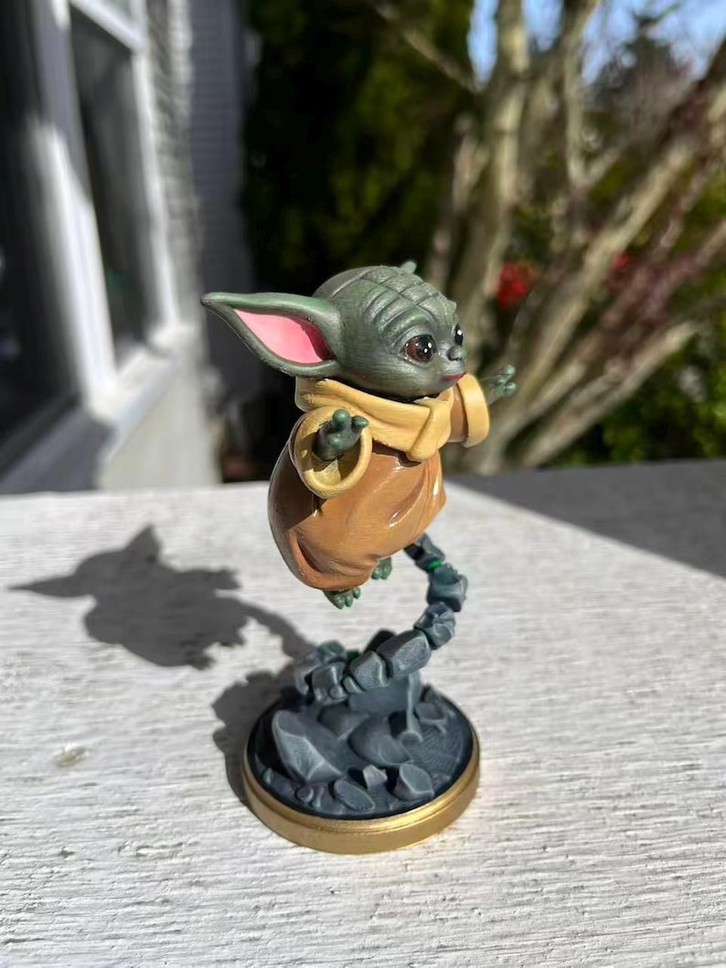 Op de afbeelding: Een beeldje van een groen alien-personage met grote oren en grote ogen, gekleed in een bruine jurk en een beige sjaal. Het beeldje zweeft in de lucht, ondersteund door een rotsachtige basis met een gouden rand. Een verzamelobject.