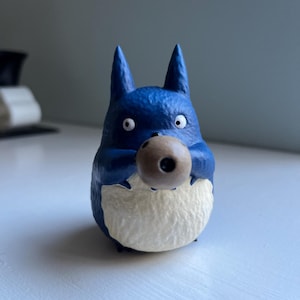 以下が含まれることがあります： 茶色の球体を持つ、青いトトロのようなキャラクターのフィギュア。白いお腹と大きな丸い目があります。フィギュアはニュートラルな背景に置かれています。