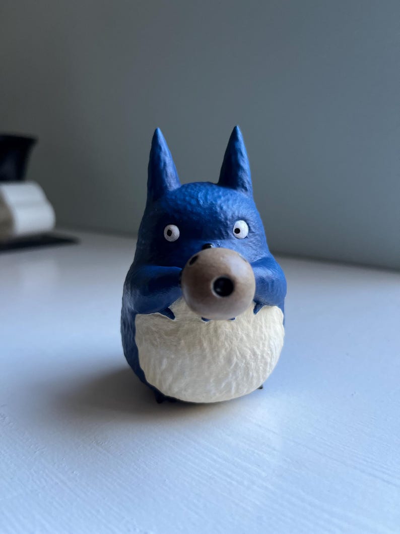 以下が含まれることがあります： 茶色の球体を持つ、青いトトロのようなキャラクターのフィギュア。白いお腹と大きな丸い目があります。フィギュアはニュートラルな背景に置かれています。