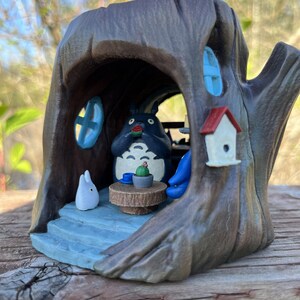 Op de afbeelding: Een handgeschilderd beeldje van Totoro in een boomstam. De sc&egrave;ne bevat een kleine tafel met een cactus, een wit konijn en een vogelhuisje. De boomstam is bruin met blauwe raamaccenten en een blauw interieur.