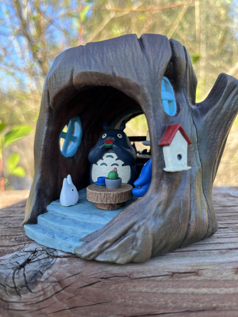 Op de afbeelding: Een handgeschilderd beeldje van Totoro in een boomstam. De sc&egrave;ne bevat een kleine tafel met een cactus, een wit konijn en een vogelhuisje. De boomstam is bruin met blauwe raamaccenten en een blauw interieur.