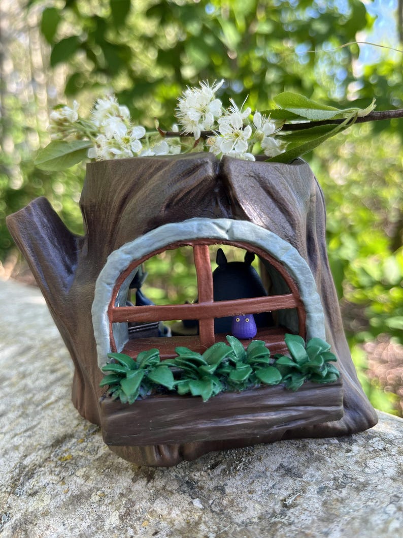 Op de afbeelding: Een bruine boomstronk-vormige plantenbak met een raam met een zwart figuur en een paars figuur. Het raam heeft een bruin frame en een groene plantenbak eronder. Witte bloemen en groene bladeren zijn in de plantenbak geplaatst.