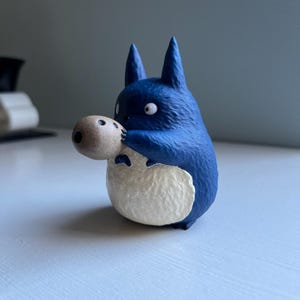 以下が含まれることがあります： 小さな丸い茶色の物体を持っている、トトロのようなキャラクターの青い置物。 このフィギュアは大きな白い目と尖った耳を持っています。 キャラクターの腹は白く、青い体と対照的です。 白い表面にあります。