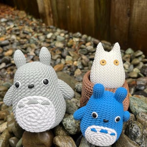 May include: Three crocheted figures resembling characters from a popular animated film. The figures are a light gray, white, and blue, with black and white facial features. They are displayed on a bed of rocks.