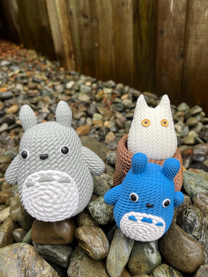 May include: Three crocheted figures resembling characters from a popular animated film. The figures are a light gray, white, and blue, with black and white facial features. They are displayed on a bed of rocks.