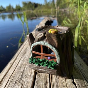 Op de afbeelding: Een grillig, bruin keramisch boomstronk huisje met een raam, groen gebladerte en fruit erop. Het miniatuurhuis staat op een verweerd houten oppervlak, met een meer en bomen op de achtergrond.
