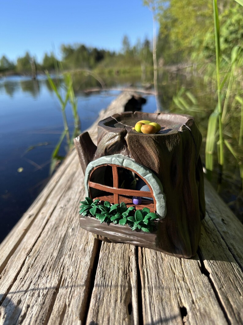 Op de afbeelding: Een grillig, bruin keramisch boomstronk huisje met een raam, groen gebladerte en fruit erop. Het miniatuurhuis staat op een verweerd houten oppervlak, met een meer en bomen op de achtergrond.