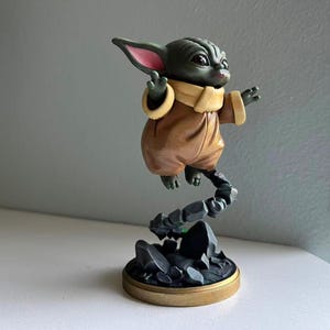 Op de afbeelding: Gedetailleerd beeldje van een personage uit een populaire sciencefictionfranchise. Het figuur is groen met grote oren, gekleed in een bruine outfit met een beige kraag. Het lijkt boven een rotsachtige basis met een goudkleurige rand te zweven.