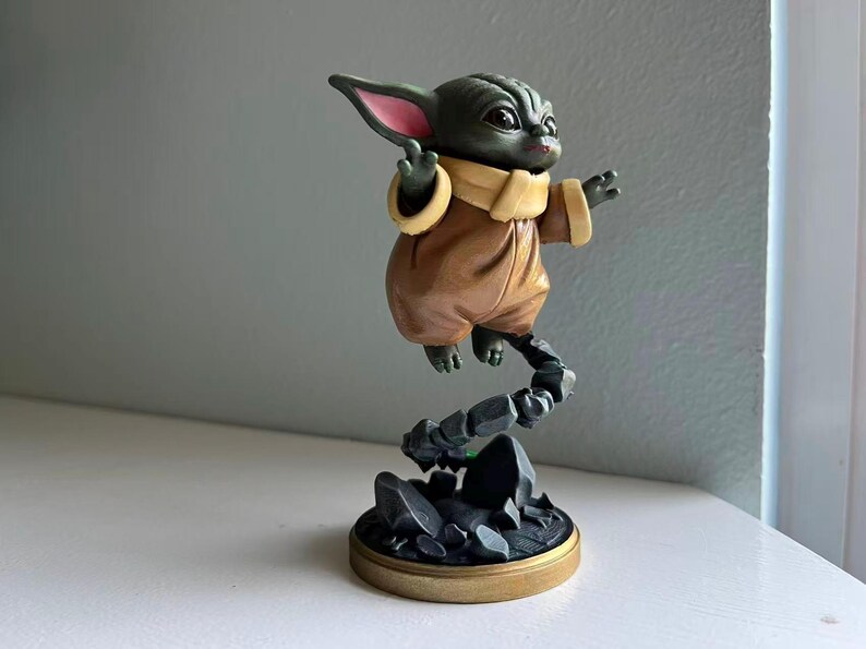 Op de afbeelding: Gedetailleerd beeldje van een personage uit een populaire sciencefictionfranchise. Het figuur is groen met grote oren, gekleed in een bruine outfit met een beige kraag. Het lijkt boven een rotsachtige basis met een goudkleurige rand te zweven.
