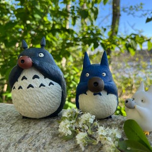 以下が含まれることがあります： アニメのキャラクター、トトロのフィギュア3体。2体は青色、1体は白色で、大きな目と丸い鼻が特徴です。フィギュアは石の表面に置かれ、背景には緑があります。