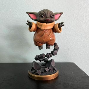 Op de afbeelding: Gedetailleerd beeldje van een personage uit een populaire sciencefiction-serie. De figuur is afgebeeld in een bruine outfit met grote oren en uitgestrekte armen, schijnbaar zwevend boven een rotsachtige basis. De basis heeft een goudkleurige rand.